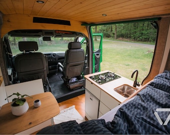 May include: A converted van interior with a small table, a plant, and a bed. The van has a kitchen area with a sink, a stovetop, and cabinets. The back door is open, revealing a view of a green grassy area.