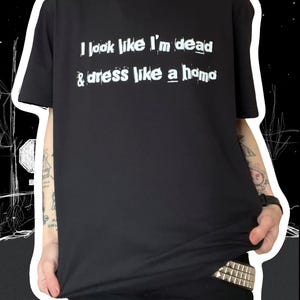 May include: Black t-shirt with the text "I look like I'm dead & dress like a homo" in white. The shirt is worn with black pants and a silver chain necklace. The style is edgy and alternative.