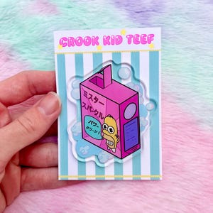 May include: A pink and purple acrylic pin featuring a cartoon character inside a juice box design. The pin has Japanese text and is on a backing card with the text "CROOK KID TEEF".