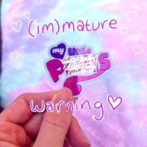 May include: A purple and pink sticker with the text "my little ponies" with a heart and a white scribble over the word "ponies". The sticker is held by a hand and the text "(im)mature warning" is written in purple below the sticker.