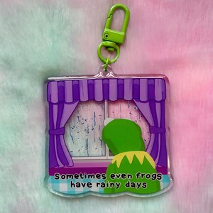 May include: A clear acrylic keyring featuring a green frog character looking out a window with purple curtains. The window shows rain and the text reads "Sometimes even frogs have rainy days."