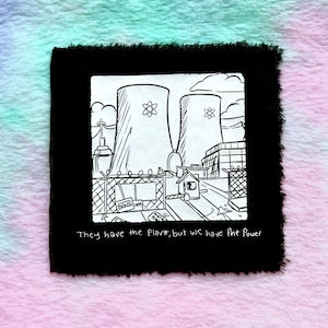 May include: A black and white illustration of a nuclear power plant on a black fabric square. The image includes two cooling towers, a fence, and the text "They have the Plant, but we have the Power."