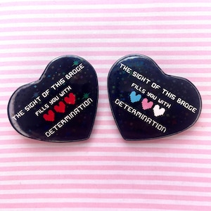 May include: Two black heart-shaped pins with a glittery finish. Each pin features the text "THE SIGHT OF THIS BROGE FILLS YOU WITH DETERMINATION" and a pixelated heart graphic in different colours.