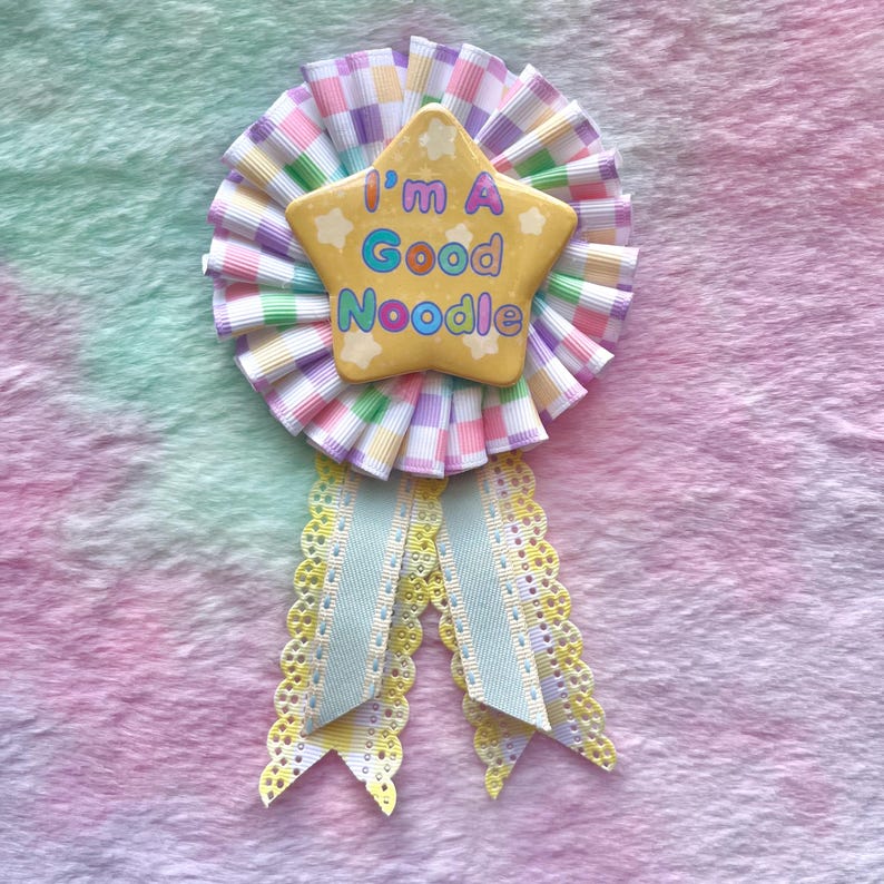 May include: A yellow star-shaped pin with the text "I'm a Good Noodle" in colorful letters. The pin is attached to a white and pastel pink checkered ribbon with a white and yellow lace ribbon.