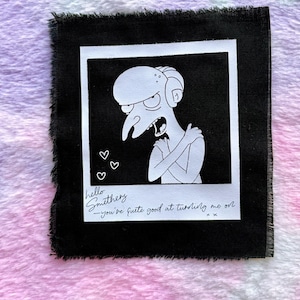 May include: A black square fabric with a white polaroid-style image of a cartoon character. The image has the text "hello Smithers - you're quite good at turning me on" with three small heart symbols. The fabric has a frayed edge.