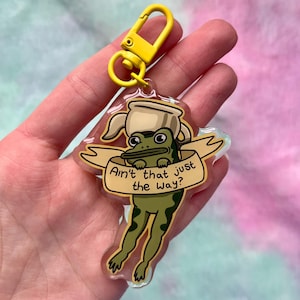 May include: A clear acrylic keychain with a cartoon frog wearing a coffee pot hat. The frog is green with dark green spots and holds a banner with the text "Ain't that just the way?" The keychain has a yellow clasp.