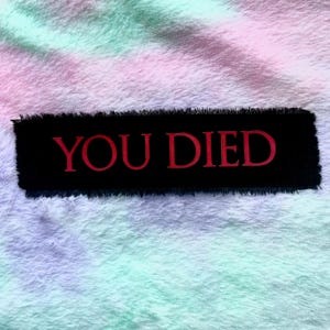May include: A black rectangular patch with the embroidered words "YOU DIED" in red. The patch has a frayed edge and is set against a soft, multi-coloured background of pastel hues.