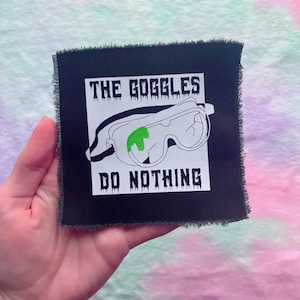 May include: Black square patch with frayed edges featuring the text "THE GOGGLES DO NOTHING" above and below a graphic of safety goggles with a green liquid. The patch is held in a hand.