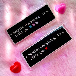 May include: Two rectangular clear acrylic keychains with the text "Despite everything, it's still you." in white. The keychains have a black background with colorful heart symbols. A pink and a red heart-shaped object are also visible.