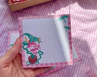 May include: Square notepad with a pink and white gingham border. The notepad features a cartoon illustration of a plant with a pink flower and green leaves. The notepad is held in a hand, with a pink striped background.