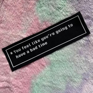 May include: Black rectangular patch with white text that reads "* You feel like you're going to have a bad time" inside a white border. The patch is on a multicolored, fluffy surface.
