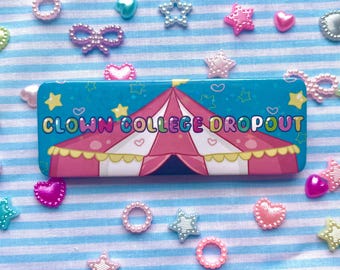May include: A blue and pink rectangular tin with a circus tent design and the text "CLOWN COLLEGE DROPOUT". The tin is surrounded by colorful plastic hearts, stars, and bows.