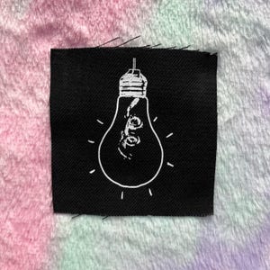 May include: A black square patch featuring a white outline of a lightbulb with a filament design inside. The lightbulb has radiating lines emanating from it. The background is a soft, multi-colored fabric.