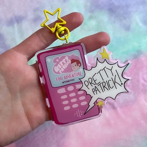 May include: A pink acrylic keychain featuring a cartoon phone design with the text "Pretty Patrick!" and "Egg Adventure Interactive!". The keychain has a yellow star-shaped clasp.