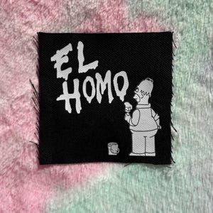 以下が含まれることがあります： 黒い正方形のパッチ。白い文字で「EL HOMO」と書かれ、漫画のキャラクターがペイントローラーを持っています。キャラクターは黒い背景に「HOMO」という言葉をペイントしています。パッチは切りっぱなしの縁です。