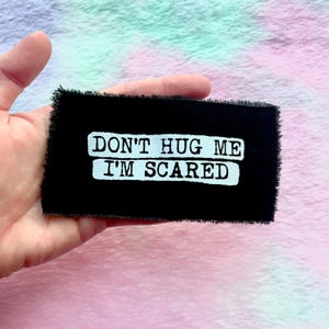 May include: A black rectangular patch with white text that reads "DON'T HUG ME I'M SCARED." The patch has a frayed edge and is set against a pastel background.