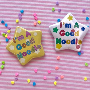 May include: Two yellow star-shaped pins with the text "I'm a Good Noodle" in rainbow letters. The pins are on a pink and white striped background with colorful sprinkles.
