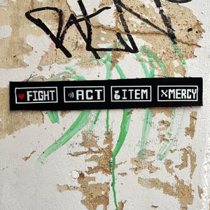 May include: A black rectangular patch with white text and borders. The text is divided into four sections: FIGHT with a heart symbol, ACT with quotation marks, ITEM with a money bag symbol, and MERCY with an X symbol. The background is a textured wall with graffiti.