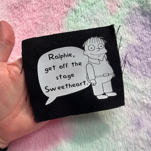 May include: Black square patch featuring a cartoon character and the text "Ralphie, get off the stage Sweetheart." The character is drawn in white, with a speech bubble containing the text. The patch has raw edges.