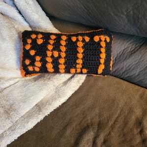 Könnte beinhalten: Ein schwarzes und orangefarbenes gehäkeltes Kissen mit dem Wort "SUI" in Orange.