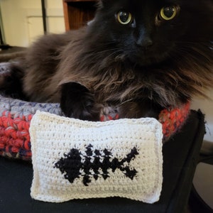 May include: A black cat with green eyes is lying in a crocheted cat bed. A white crocheted pillow with a black fish design is in front of the cat.
