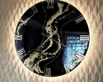 Peut inclure: Une horloge murale ronde avec un cadran noir et des chiffres romains argentés. Le cadran présente un motif marbré or et blanc. L'horloge est éclairée d'une lumière douce et montée sur un mur texturé.