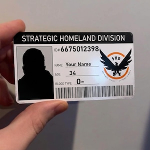 Puede incluir: Una tarjeta de identificación rectangular con el texto "STRATEGIC HOMELAND DIVISION" en la parte superior. La tarjeta presenta una silueta, el número de identificación 6675012398 y el nombre "Your Name". También incluye la edad "34", el grupo sanguíneo "0-" y un emblema con un pájaro.