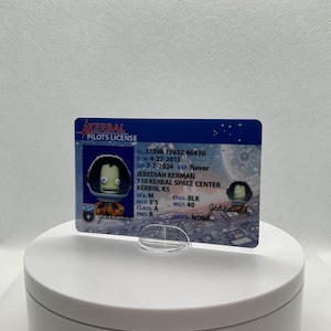 May include: A Kerbal Pilots License card featuring Jebediah Kerman. The card has a blue background with white text and a cartoon image of a Kerbal character. The card includes personal details such as date of birth, height (3'5), and weight (40).