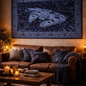 May include: A large, dark blue tapestry depicting a spaceship against a starburst design, framed with an ornate border. It hangs above a brown leather sofa with cushions and a throw. A wooden coffee table and candles create a warm, inviting living room setting.