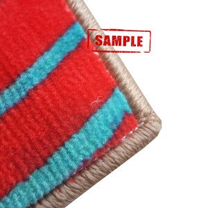 Pu&ograve; includere: Primo piano del bordo di un tappeto a strisce rosse e turchesi con un bordo beige. Il tappeto ha una trama tessuta ed &egrave; etichettato "SAMPLE".