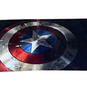 Pu&ograve; includere: Un primo piano dello scudo di Capitan America, con un design rosso, bianco e blu. Lo scudo &egrave; fatto di metallo e ha un aspetto usurato e invecchiato.