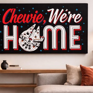 May include: A black canvas wall art piece with the text "Chewie, We're HOME" in red and white lettering. The artwork features a Millennium Falcon design. The art is displayed above a sofa with pillows and a wooden table.