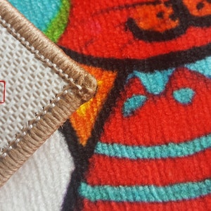 Pu&ograve; includere: Primo piano di un tappeto beige con un bordo marrone. Il tappeto ha un retro bianco con un motivo a griglia e la parola "SAMPLE" stampata in rosso. Il tappeto &egrave; piegato per rivelare un motivo colorato con accenti rossi, blu e gialli.