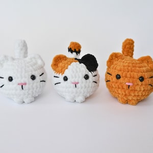 May include: Three crocheted cat plushies. One is white, one is white with black and orange patches, and one is orange. All have black eyes and pink noses.