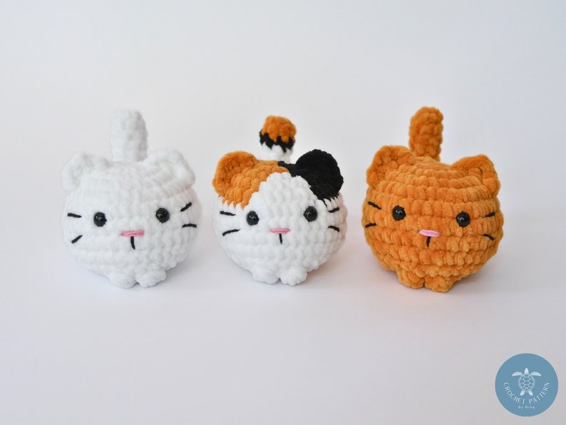 May include: Three crocheted cat plushies. One is white, one is white with black and orange patches, and one is orange. All have black eyes and pink noses.