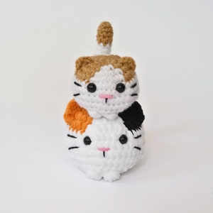 May include: Two crocheted cat plushies stacked on top of each other. The top cat is brown, white, and orange, and the bottom cat is white with black and orange accents. Both cats have black eyes and pink noses.
