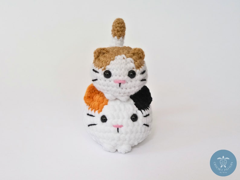 May include: Two crocheted cat plushies stacked on top of each other. The top cat is brown, white, and orange, and the bottom cat is white with black and orange accents. Both cats have black eyes and pink noses.