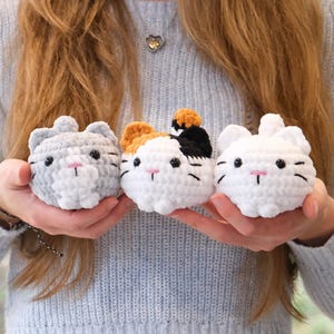 May include: Three crocheted cat plushies in different colors and patterns. The cat on the left is gray with white accents, the middle cat is white with orange, black, and white accents, and the cat on the right is white.