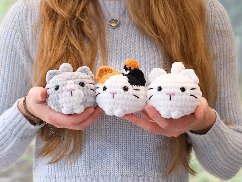 May include: Three crocheted cat plushies in different colors and patterns. The cat on the left is gray with white accents, the middle cat is white with orange, black, and white accents, and the cat on the right is white.