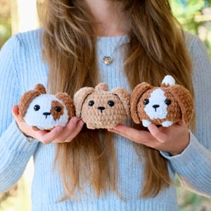 Pu&ograve; includere: Tre giocattoli di peluche per cani all'uncinetto. I cani sono marroni e bianchi con occhi e nasi neri. I cani sono seduti nelle mani di una persona.