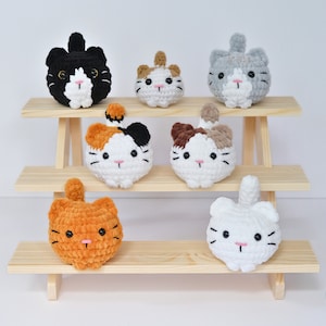May include: Seven crocheted cat plushies in various colors and patterns, displayed on a wooden shelf. The plushies are all round and have a cute, cartoonish appearance.