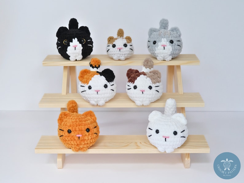 May include: Seven crocheted cat plushies in various colors and patterns, displayed on a wooden shelf. The plushies are all round and have a cute, cartoonish appearance.