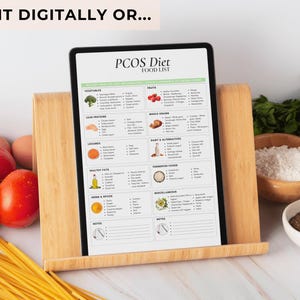 May include: A printable PCOS diet food list on a tablet propped up on a wooden stand. The list includes categories for vegetables, lean proteins, legumes, healthy fats, herbs & spices, fruits, whole grains, dairy & alternatives, fermented foods, and miscellaneous. The tablet is surrounded by various food items, including tomatoes, pasta, rice, and spices.