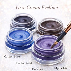 May include: Four jars of Luxe Cream Eyeliner in various shades: Carbon Luxe, Electric Petal, Dark Roast, and Mystic Iris. A makeup brush is poised above the Mystic Iris jar. The jars are clear with silver lids, set against a sparkling background.