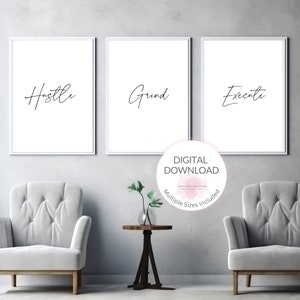 May include: Three white framed prints with black calligraphy on a white background. The prints read "Hustle", "Grind", and "Execute". The prints are hanging on a gray wall above two gray armchairs and a small table with a potted plant.