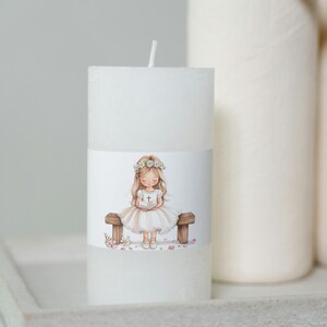 May include: White candle with a printed image of a girl in a white dress and a flower crown sitting on a bench holding a book. The girl is surrounded by pink flower petals.