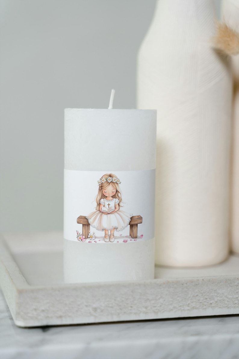 May include: White candle with a printed image of a girl in a white dress and a flower crown sitting on a bench holding a book. The girl is surrounded by pink flower petals.
