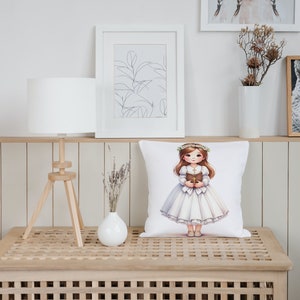 May include: White pillow with a printed image of a girl in a white dress holding a book. The girl is wearing a flower crown and has brown hair. The pillow is on a wooden table with a lamp and a vase of flowers.