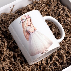 May include: White ceramic mug with a colorful illustration of a girl wearing a white dress and a flower crown. The girl is holding her hands together in a prayer position.