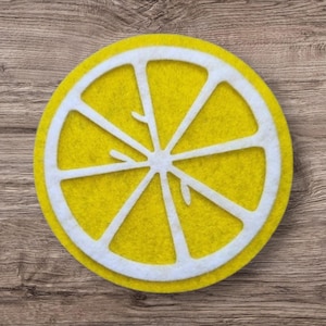 May include: A circular felt coaster shaped like a lemon slice, featuring a bright yellow background with a white felt overlay depicting the segmented structure of a citrus fruit, including a central core and radiating wedge-shaped sections.